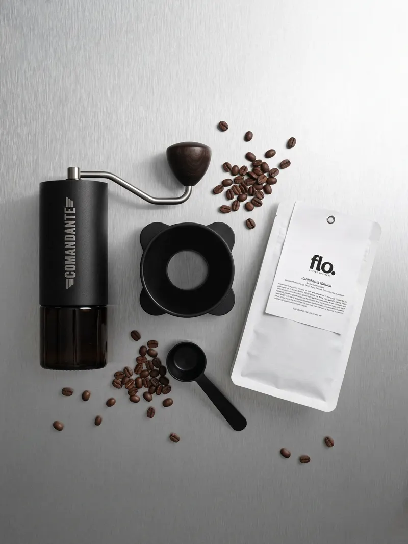 A hand grinder, scoop and roasted beans beside a flo. coffee pouch on brushed steel.